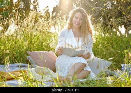 Ganzkörper wunderschöne Inhalte Frau mit Hintergrundbeleuchtung trägt ein stilvolles Sonnenkleid, liest Buch und sieht mit einem Lächeln auf die Kamera, während sie sich auf einer üppigen Wiese in s ausruht Stockfoto