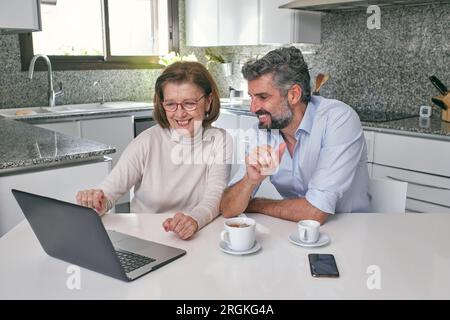 Lächelnde ältere Frau, die auf den Laptop zeigte, während sie mit einem Mann mittleren Alters am Tisch saß, mit Bart und Tassen mit Getränken in der modernen Marmorküche zu Hause Stockfoto