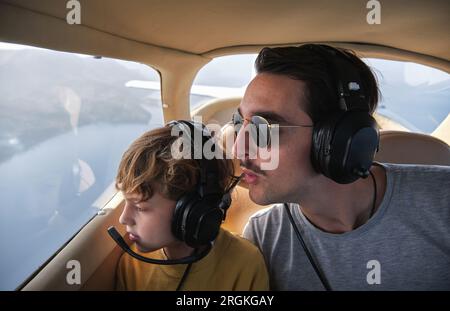 Junger Mann und Sohn mit Kopfhörern fliegen im Flugzeug über dem Meer und schauen mit Interesse aus dem Fenster Stockfoto