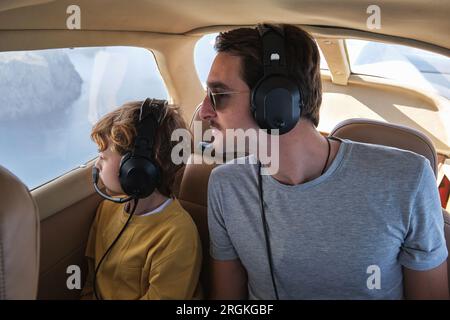 Junger Mann und Sohn in legerer Kleidung und Kopfhörern fliegen im Flugzeug und schauen neugierig aus dem Fenster während der Reise Stockfoto