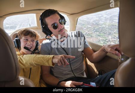 Aufgeregter kleiner Junge, der in lässiger Kleidung lächelt und mit einem Headset lächelt und wegzeigt, während er mit dem jungen Vater während des Ausflugs in der Flugzeugkabine sitzt Stockfoto