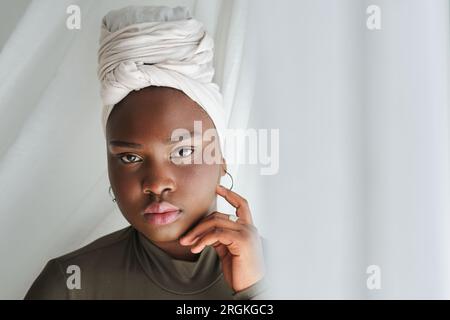 Porträt einer attraktiven, emotionslosen afroamerikanischen Frau im Turban, die einen lässigen Pullover und Ohrringe trägt und das Gesicht berührt, während sie in die Kamera schaut Stockfoto