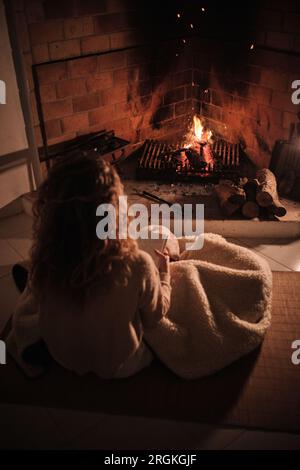 Rückansicht einer jungen Frau, die sich auf dem Boden neben einem brennenden Kamin ruht und ein Buch im gemütlichen Wohnzimmer zu Hause liest Stockfoto