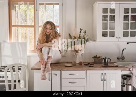 Der ganze Körper einer jungen Frau in legerer Kleidung sitzt an Küchenmöbeln und lächelt in der gemütlichen Küche zu Hause Stockfoto