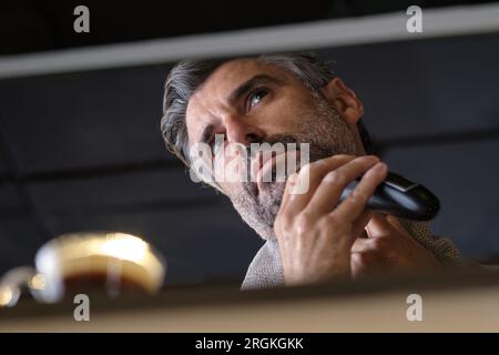 Von unten: Ein konzentrierter Mann mit grauen Haaren, der morgens Routine macht und sich zu Hause mit dem Trimmer rasiert Stockfoto