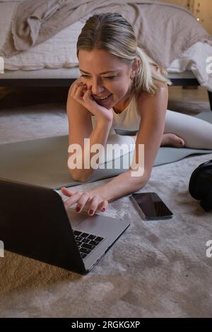 Lächelnde Sportlerin in Sportbekleidung sitzt auf der Matte und sucht nach einem Online-Tutorial, während sie sich auf den Yoga-Kurs zuhause im Schlafzimmer vorbereitet Stockfoto