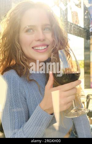 Porträt einer schönen, positiven, jungen, lockigen Frau mit einem Glas Rotwein, die in die Kamera schaut Stockfoto