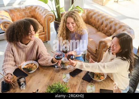 Ein großer Winkel von jungen, multirassischen Freunden, die mit Essen und Wein am Tisch sitzen und lachen Stockfoto