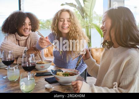 Lachende Freunde in lässiger Kleidung, Spaß beim Essen von Ramen und beim Trinken von Wein im Restaurant Stockfoto