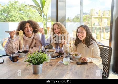 Eine Gruppe verschiedener Freunde, die sich mit leckeren Ramen mit Stäbchen verbinden und essen, während sie in der Nähe des Fensters im modernen Restaurant sitzen Stockfoto