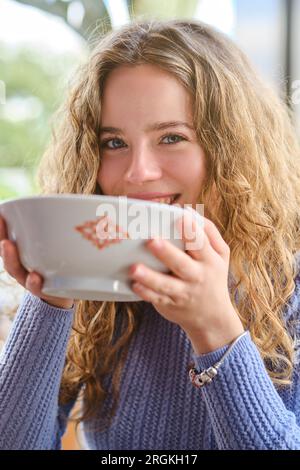 Charmante Frau mit lockigem Haar in Strickpullover, die in die Kamera schaut und Ramen-Suppe aus Keramikschüssel trinkt, während sie Zeit in einem asiatischen Restaurant verbringt Stockfoto