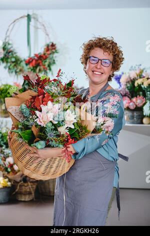 Positiver weiblicher Florist in der Schürze mit einem Haufen frischer Blumen im Korb, der auf die Kamera schaut, während er in einem Blumenladen arbeitet Stockfoto
