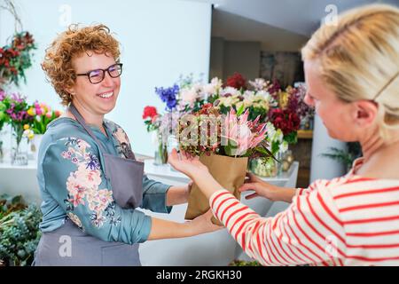 Positiver weiblicher Florist in Gläsern und Schürze, der dem Kunden während des Arbeitstages in Blumenform ein Bouquet aus frischen Blütenhornsirup und protea in Papiertüte gibt Stockfoto