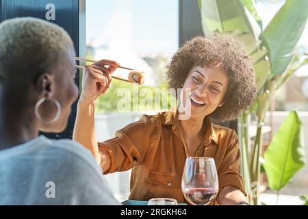 Lächelnde junge ethnische Frau mit lockigem Haar, die im Stuhl sitzt und während des Meetings köstliches Sushi isst Stockfoto