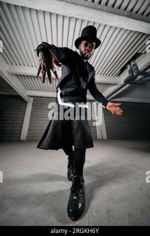 Der ganze Körper des gefühlslosen afroamerikanischen Mannes mit Accessoires in extravagantem schwarzen Outfit und einem großen Hut, der in Garage n tanzt Stockfoto
