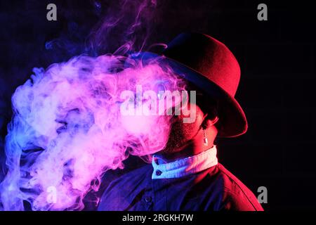 Seitenansicht des jungen schwarzen Mannes in stilvollem Outfit und Hut, der in dunklem Raum mit Rauch steht Stockfoto