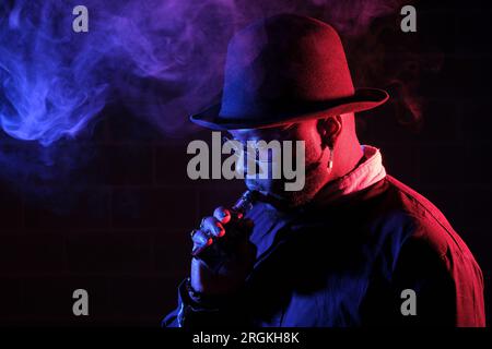 Seitenansicht eines bärtigen Mannes in einem Hut, der Vape raucht, während er mit Sonnenbrille in einem dunklen Raum steht Stockfoto