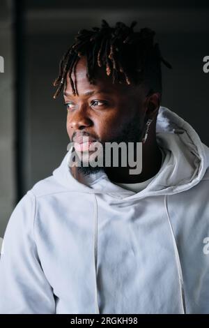 Seriöses männliches Modell in trendigen Hoodie und mit Dreadlocks, die auf verschwommenem Hintergrund stehen Stockfoto