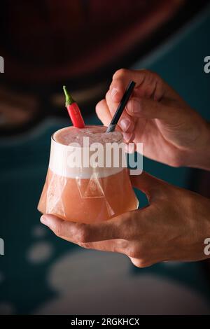 Eine Person, die köstliche Cocktails mit Chili-Pfeffer vorführt und in Glas mit Stroh auf verschwommenem Hintergrund serviert wird Stockfoto