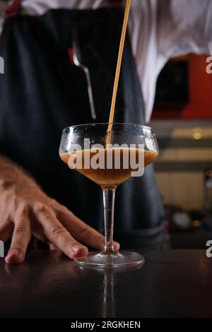 Der anonyme Barkeeper in Uniform steht neben der Theke und gießt während des Arbeitstages in der Bar einen frischen Kaffee-Cocktail in ein Champagner-Coupé Stockfoto