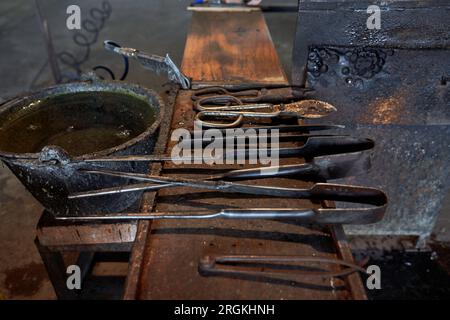 Metallinstrument für die Arbeit mit heißem Glas Stellen auf Holztisch in der Nähe von Keramiktopf in dunklen Gebäuden mit verschwommenem Hintergrund Stockfoto