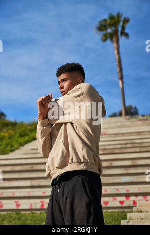 Sportlicher hispanischer Männchen in Activewear, der sich vor dem Workout aufwärmt, während er mit einem Baum auf den Stufen der sonnigen Straße der Stadt steht Stockfoto