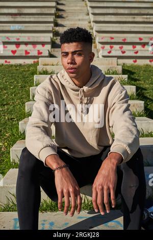 Seriöser hispanischer Mann mit Hoodie und Leggings, der auf Treppen neben dem Skateboard sitzt, um Hobby zu üben Stockfoto