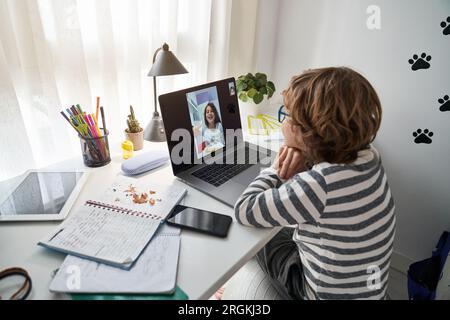 Seitenansicht eines jungen Kindes mit lockigem Haar, der am Tisch sitzt und sich mit einem Freund per Netbook per Video chatten lässt, während er Hausaufgaben im Zimmer macht Stockfoto