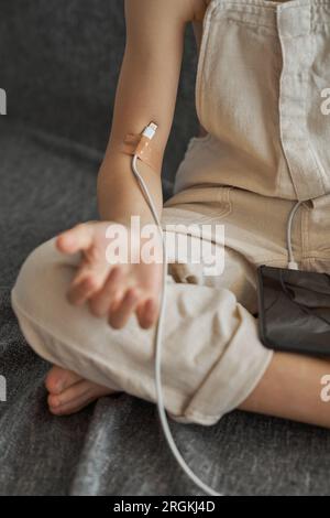 Nicht erkennbares, digital abhängiges Kind mit Ladekabel, das mit dem Arm verbunden ist, mit Pflaster, das auf dem Boden sitzt und mit Smartphone an grauem Hintergrund anliegt Stockfoto