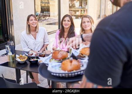 Der männliche Kellner trägt ein Tablett mit Burgern, serviert einer Gruppe positiver, lächelnder Freundinnen Essen und isst zusammen im Straßencafé zu Mittag Stockfoto