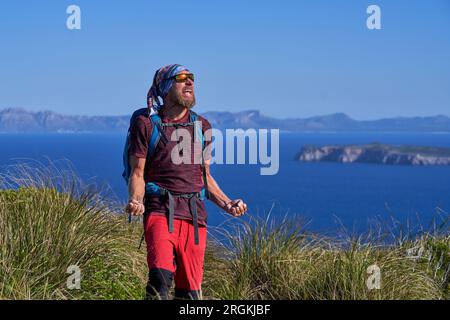 Aufgeregter männlicher Wanderer in Sonnenbrille mit Rucksack, der auf einem grünen Grashügel vor dem blauen See in den Bergen steht und mit geballten Fäusten schreit, während Cel Stockfoto