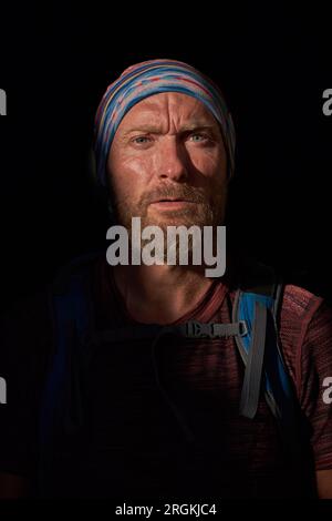 Nachdenklicher, bärtiger Reisender in Bandana mit runzelnden Augen und Blick auf die Kamera, während er im Schatten mit Sonnenlicht steht Stockfoto