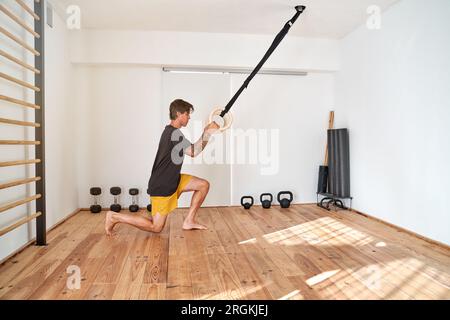 Barfußläufer männlicher Sportler in Sportbekleidung steht auf dem Knie und trainiert mit hölzernen Turnringen während intensivem Training im Fitnessstudio Stockfoto