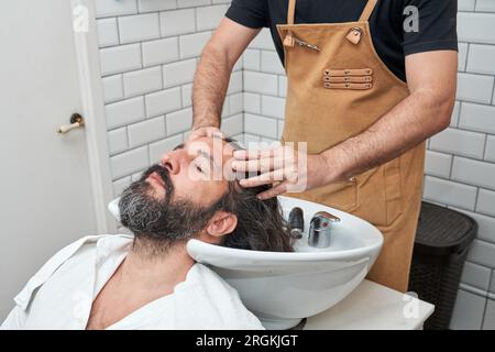 Anonymer Barbier in der Schürze, der lange Haare von Mann mit Shampoo in weißer Spüle wäscht, im professionellen Friseursalon Stockfoto