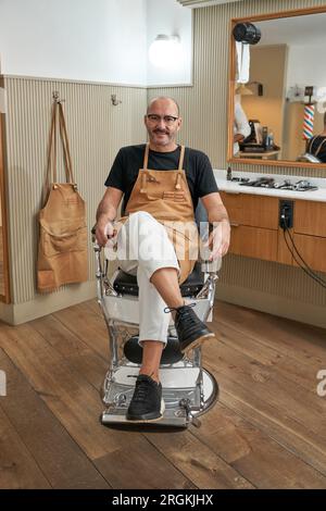 Der ganze Körper eines bärtigen Barbiers in der Schürze sitzt auf einem Stuhl und schaut in die Kamera, während er eine Pause im Friseursalon macht Stockfoto