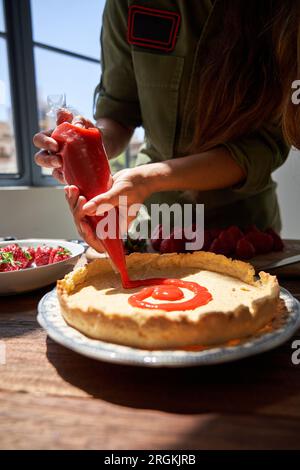 Schneide die weibliche Köchin in lässiger Kleidung, die am Tisch steht, und lege Beerensauce auf die Kuchenkruste, während du leckeren Kuchen zubereitest Stockfoto