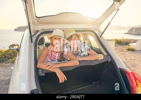 Bruder umarmt seine Schwester, während sie lächelt und Stroh trägt Hüte in den Rücksitzen eines Autos kleben ihre Kopf aus dem Kofferraum mit dem Meer auf Stockfoto