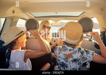 Blonde Mutter mit Sonnenbrille drehen in den Sitz eines Auto und Blick auf ihre Kinder sitzen im Rücken Sitze, die Strohhüte tragen und tun Stockfoto