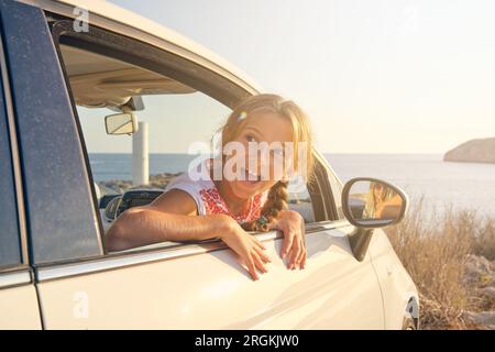 Blonde Mädchen stecken ihren Kopf aus dem Fenster von Ein Auto vor dem Meer mit einem geparkten Glücklicher Ausdruck und die Reflexion ihrer Mutter in der Seite mi Stockfoto