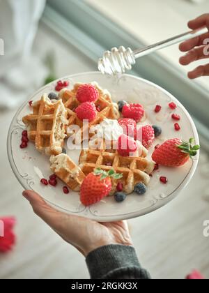 Eine anonyme Person von oben gießt Honig auf süße gebackene Waffeln mit reifen Beeren, serviert auf einem Tisch in der Küche Stockfoto