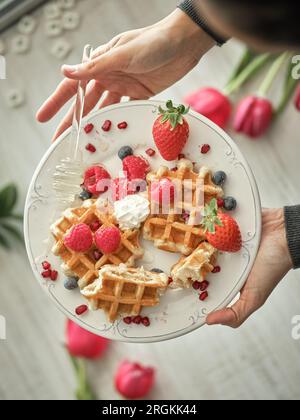 Eine anonyme Person von oben gießt Honig auf süße gebackene Waffeln mit reifen Beeren, serviert auf einem Tisch in der Küche Stockfoto