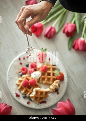 Eine anonyme Person von oben gießt Honig auf süße gebackene Waffeln mit reifen Beeren, serviert auf einem Tisch in der Küche Stockfoto