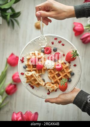 Eine anonyme Person von oben gießt Honig auf süße gebackene Waffeln mit reifen Beeren, serviert auf einem Tisch in der Küche Stockfoto