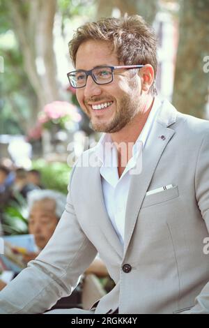 Optimistischer junger Mann im formellen Anzug mit Brille, der im Park vor verschwommenem Hintergrund sitzt, glücklich lächelt und wegschaut Stockfoto