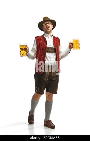 Junger attraktiver Schnurrbart mit Hut, trägt bayerisches Outfit, hält Bierbecher und singt auf weißem Hintergrund. Oktoberfest-Konzept Stockfoto