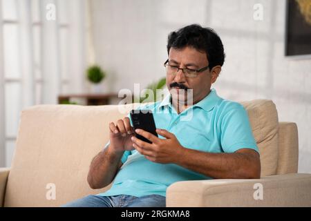 Indischer Mann mittleren Alters mit Brille beschäftigt mit Handy beim Sitzen auf dem Sofa zu Hause - Konzept von Digital Engagement, Social Media und Stockfoto