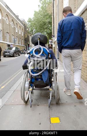 Rückansicht Mann, der mit behinderter Person im Rollstuhl und Atemröhren in London England, Großbritannien KATHY DEWITT Stockfoto