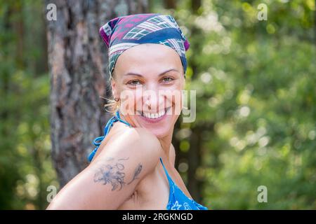 40 Jahre alte europäische Frau im Kopftuch und Badeanzug lächelt freundlich in die Kamera. Tourist im Wald im Urlaub Stockfoto