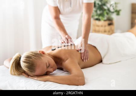 Eine wunderschöne Frau genießt eine entspannende Rückenmassage im kosmetischen Wellnesscenter. Die Masseurin trägt Lavasteine auf die Wirbelsäule auf. Stockfoto