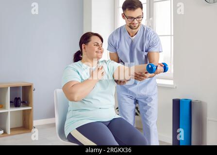 Der Arzt hilft einer glücklichen, übergewichtigen Patientin, Physiotherapietraining mit Kurzhanteln zu machen Stockfoto